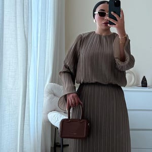 以下が含まれることがあります： プリーツ加工が施されたオリーブブラウンのドレス。長袖と、お揃いのスカート。小さな茶色のハンドバッグがドレスの前に持たれています。サングラスと黒いヘッドカバーが組み合わされています。
