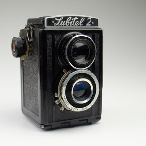 Op de afbeelding: Een zwarte Lubitel 2-tweelinglensreflexcamera met een zilveren naamplaatje en twee lenzen. De camera heeft een zwarte kunstlederen bekleding en een zilveren opwindknop.