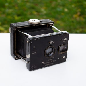 May include: A black Tenax folding camera with a C.P. Goerz lens. The camera has a black leather covering and a silver metal lens. The camera is in good condition and has a vintage look.