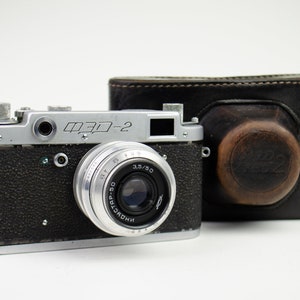Puede incluir: Una cámara de película de 35 mm negra y plateada con una funda de cuero. La cámara tiene una lente etiquetada como "FED-2" y "3.5/50".