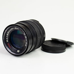 Op de afbeelding: Een zwarte camera lens met de tekst "Калейнар-5Н" en "МО" in Cyrillisch schrift. De lens heeft een diafragma van 2.8/100 mm. De lens wordt geleverd met twee zwarte lensdoppen.