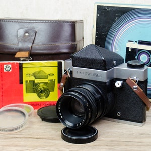 May include: A black and silver Kiev 60 medium format film camera with a black lens and a brown leather strap. The camera is sitting on a wooden surface with a brown leather case, a film canister, and several lens filters.