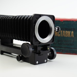 Peut inclure: Un tube d'extension d'appareil photo à soufflet noir avec une bague argentée et deux boutons noirs. Le tube est fixé à une base métallique noire avec un bouton argenté. Le texte "Vorsatz Mctabka Dispositif Attachment" est visible sur une boîte rouge et bleue en arrière-plan.