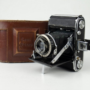 May include: A black and silver folding camera with a leather case. The camera has a lens with the text "COMPUR-RAPID" and "1:4.5 f=75mm" printed on it. The case has the text "Semi-Princes" printed on it.