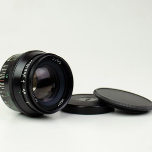 May include: A black camera lens with a 50mm focal length and a maximum aperture of f/2.0. The lens has a series of measurement markings and is accompanied by two black lens caps. The lens is on a white surface.