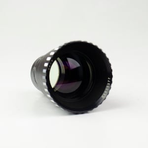 Op de afbeelding: Een zwarte camera lens met een zilveren ring rond de rand. De lens ligt op een wit oppervlak.