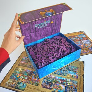 Pode incluir: Uma caixa de madeira roxa com uma tampa azul e dourada, aberta para revelar um quebra-cabe&ccedil;a de madeira com pe&ccedil;as intrincadas. A caixa tem o texto "Dreamion Puzzles" na frente. O quebra-cabe&ccedil;a &eacute; uma cena de Natal com uma variedade de edif&iacute;cios e personagens.