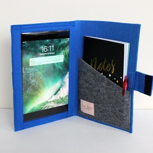 Op de afbeelding: Een blauwe felt tablet hoes met een grijze felt zak en een zwart notitieboekje met de tekst "Notes" in gouden letters. Een rode pen steekt uit de zak.