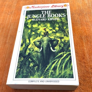 May include: A white book cover with a green and brown illustration of a man riding an elephant through a jungle. The title is "The Jungle Books" by Rudyard Kipling. The book is part of the "Masterpiece Library" series and is complete and unabridged.
