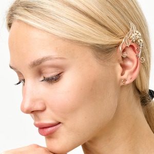 Op de afbeelding: Een goudkleurige oor manchet met bloemen- en bladeren ontwerpen. De manchet wikkelt zich om het oor en heeft delicate, wervelende details. De sieraden worden gedragen op het oor van een persoon met blond haar.