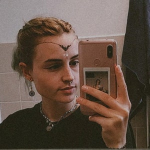 May include: A person with short, light blonde hair is taking a selfie in a bathroom mirror. They are wearing a black shirt, a silver necklace with a pendant, and a silver nose ring. They have a silver and black headpiece with a crystal hanging from it. The person is holding a pink smartphone with a photo of a person in a vintage dress inside the phone case.