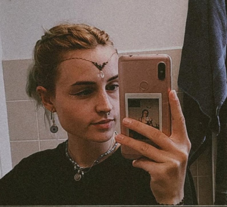 May include: A person with short, light blonde hair is taking a selfie in a bathroom mirror. They are wearing a black shirt, a silver necklace with a pendant, and a silver nose ring. They have a silver and black headpiece with a crystal hanging from it. The person is holding a pink smartphone with a photo of a person in a vintage dress inside the phone case.