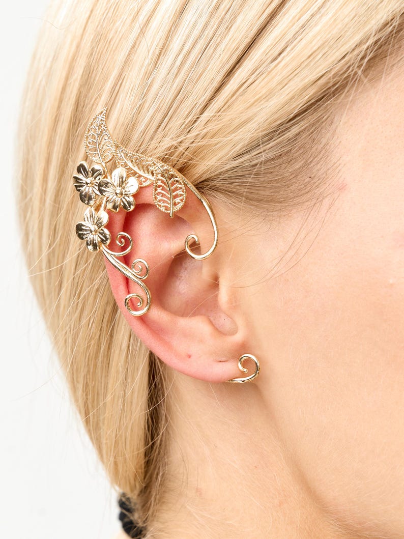 Op de afbeelding: Goudkleurige oor manchet met bloemen- en bladerenmotieven. De manchet omsluit het oor en bevat een kleine oorbel. De sieraden worden gedragen op het oor van een persoon met blond haar.