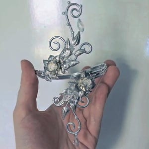 May include: A silver-toned metal arm cuff featuring intricate wire-wrapped scrollwork, metallic leaves, and clear faceted crystal accents. The design includes two floral-shaped centerpieces and two dangling teardrop-shaped clear crystals.