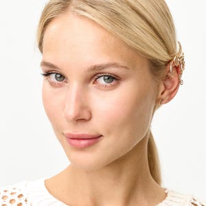 Op de afbeelding: Een vrouw met blond haar draagt een gouden oormanchet en kleine gouden oorringen. De oormanchet heeft een decoratief ontwerp dat rond het oor buigt. Ze draagt een witte, opengebreide trui.