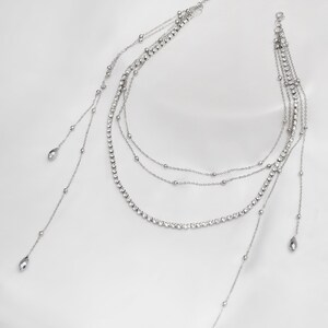 May include: A silver layered necklace with a delicate chain and crystal accents. The necklace has three strands with a longer strand on each side that ends with a teardrop-shaped pendant.