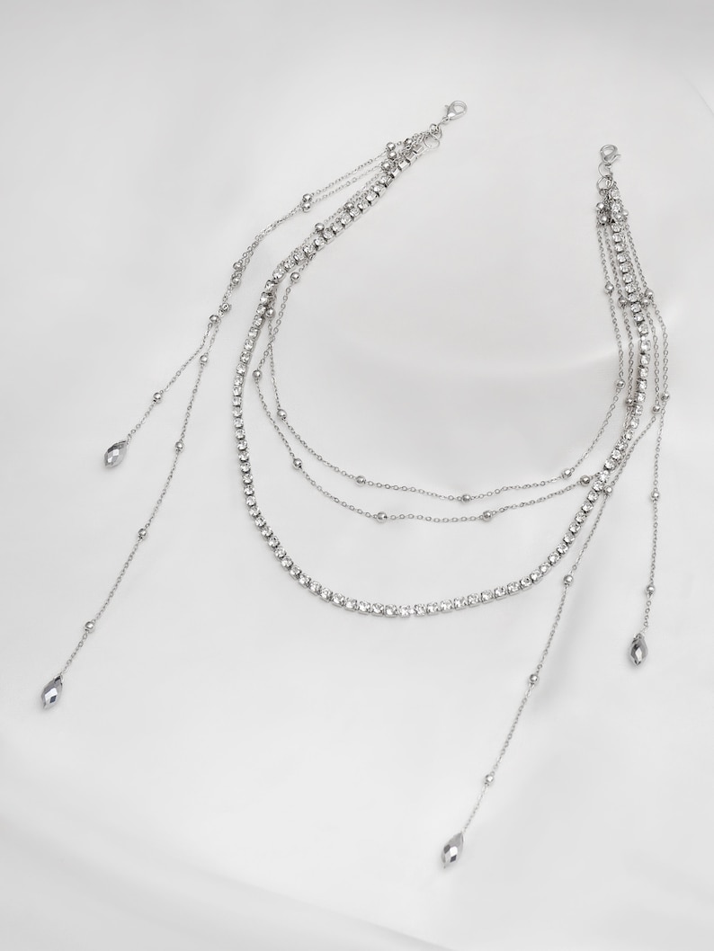 May include: A silver layered necklace with a delicate chain and crystal accents. The necklace has three strands with a longer strand on each side that ends with a teardrop-shaped pendant.
