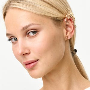 Op de afbeelding: Een goudkleurige oor manchet met een bloemen- en bladeren ontwerp. De oor manchet wikkelt zich om het oor en wordt vastgezet met een kleine haak. De sieraden worden gedragen op het oor van een persoon met blond haar.