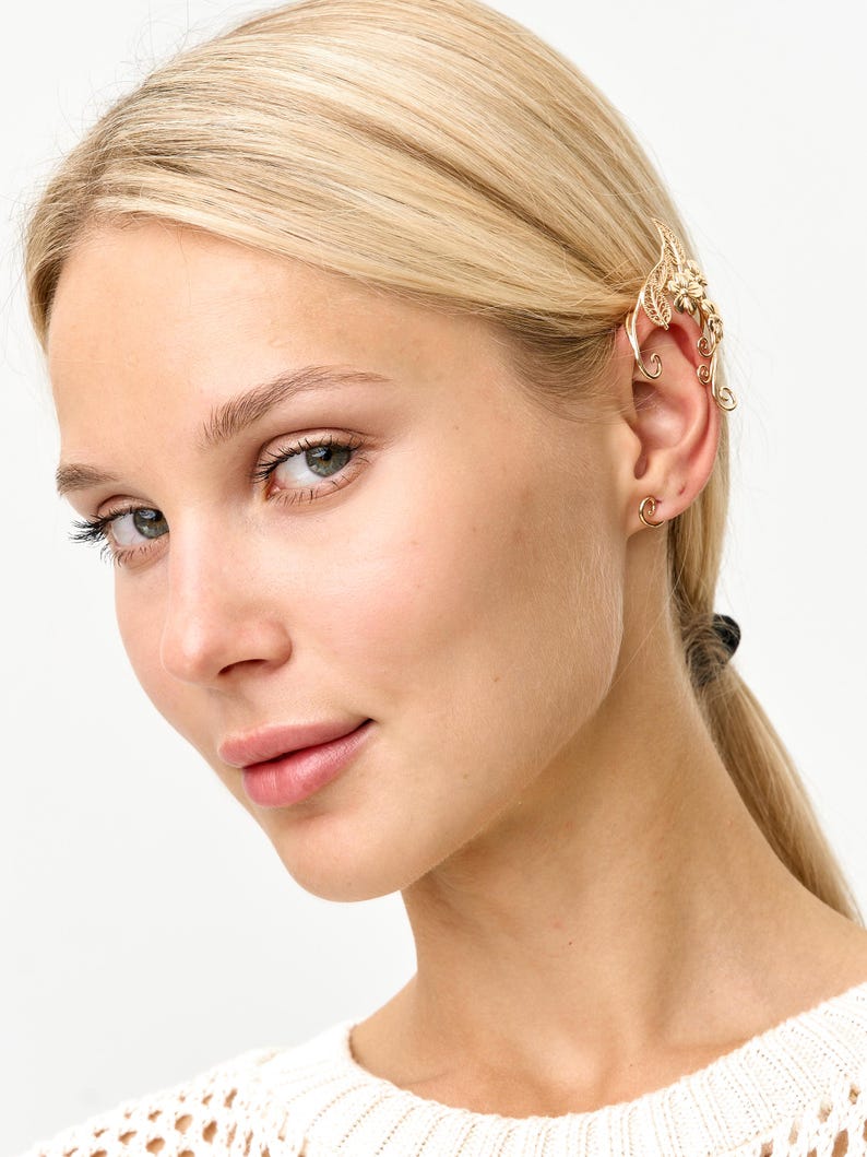 Op de afbeelding: Een goudkleurige oor manchet met een bloemen- en bladeren ontwerp. De oor manchet wikkelt zich om het oor en wordt vastgezet met een kleine haak. De sieraden worden gedragen op het oor van een persoon met blond haar.