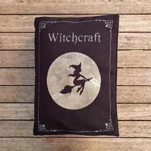 May include: A black pillow with a white moon and a witch silhouette flying on a broomstick. The pillow has the word "Witchcraft" written in white on the top.