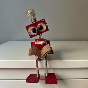 Puede incluir: Un robot de madera rojo con una cabeza de bombilla y grandes ojos redondos est&aacute; leyendo un libro. El robot tiene un cuerpo de metal y est&aacute; sentado sobre una superficie blanca.