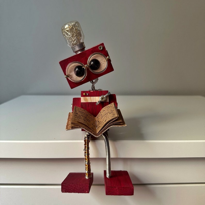 Puede incluir: Un robot de madera rojo con una cabeza de bombilla y grandes ojos redondos est&aacute; leyendo un libro. El robot tiene un cuerpo de metal y est&aacute; sentado sobre una superficie blanca.