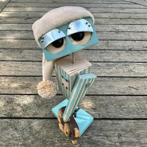 May include: A whimsical wooden robot with a blue and white striped body, a white fuzzy hat, and blue eyes. The robot is holding a wooden stick with a blue and white striped design.