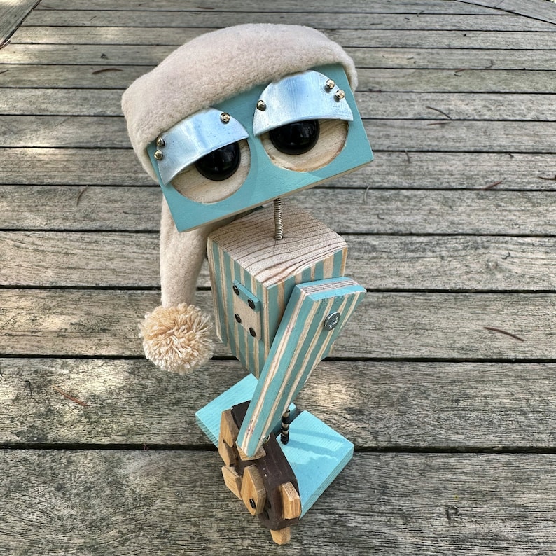 May include: A whimsical wooden robot with a blue and white striped body, a white fuzzy hat, and blue eyes. The robot is holding a wooden stick with a blue and white striped design.