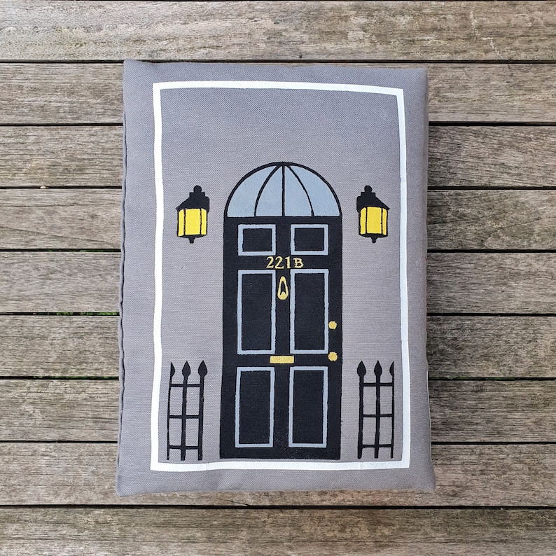 May include: A gray and white pillow with a black door with the number 221B on it. The door has two windows and a gold door knocker. There are two yellow lights on either side of the door.