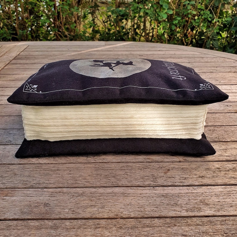 May include: A black and white book-shaped pillow with a moon and a witch silhouette on the front. The pillow is made of a soft, plush fabric and has a white, fluffy filling.
