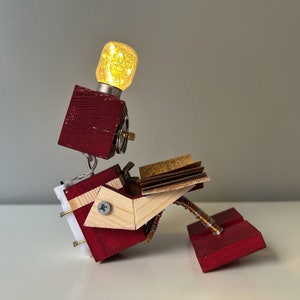 Puede incluir: Una escultura de robot de madera con una cabeza de bombilla, leyendo un libro. El robot est&aacute; hecho de madera roja y natural y tiene un brazo de alambre de cobre.