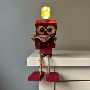 Puede incluir: Una figurita de robot de madera con una cabeza de bombilla, que lleva gafas y lee un libro. El robot est&aacute; sentado en una superficie blanca con las piernas colgando sobre el borde.