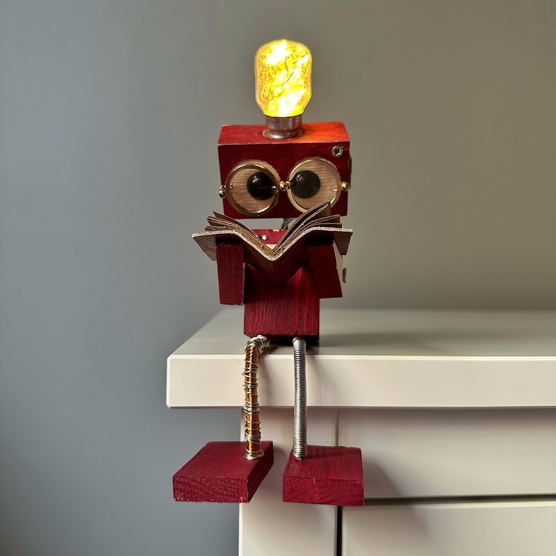 Puede incluir: Una figurita de robot de madera con una cabeza de bombilla, que lleva gafas y lee un libro. El robot est&aacute; sentado en una superficie blanca con las piernas colgando sobre el borde.