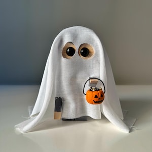May include: A white ghost figurine with large black eyes, holding a small orange pumpkin with a carved jack-o'-lantern face. The ghost is wearing a white sheet and has a wooden body.