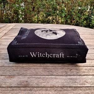 May include: A black book-shaped pillow with the word "Witchcraft" printed in silver on the front. The pillow features a silver silhouette of a witch flying on a broomstick in front of a full moon.
