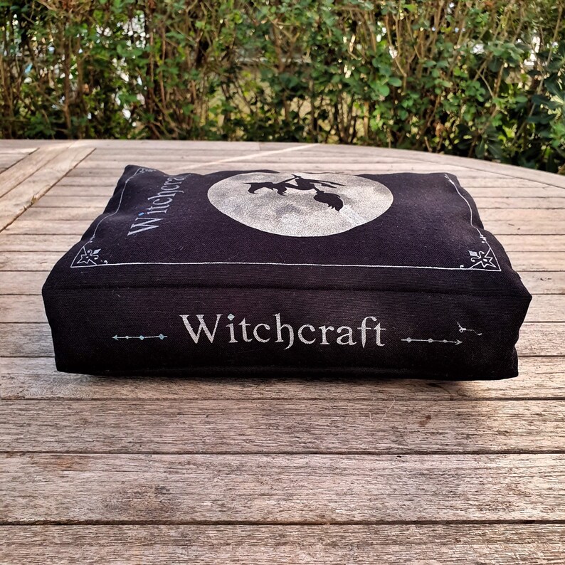 May include: A black book-shaped pillow with the word "Witchcraft" printed in silver on the front. The pillow features a silver silhouette of a witch flying on a broomstick in front of a full moon.