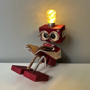 Puede incluir: Un robot de madera rojo con una bombilla en la cabeza, que lleva gafas y lee un libro. El robot est&aacute; sentado sobre una superficie blanca.