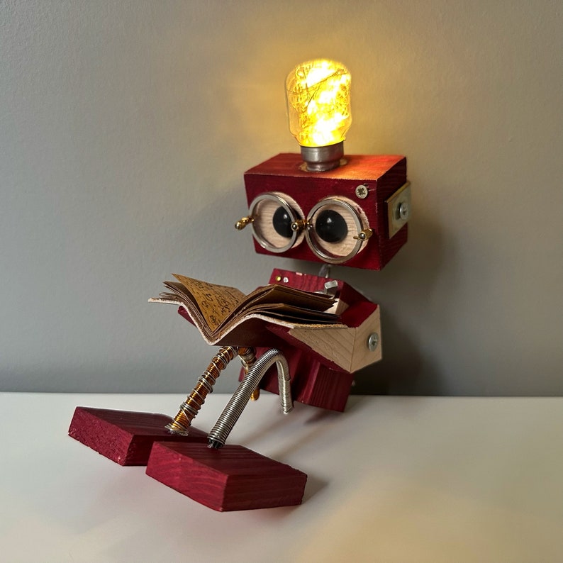 Puede incluir: Un robot de madera rojo con una bombilla en la cabeza, que lleva gafas y lee un libro. El robot est&aacute; sentado sobre una superficie blanca.