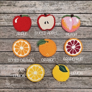May include: Nine felt fruit coasters, including a whole apple, a sliced apple, a peach, a whole orange, a sliced orange, a grapefruit, a whole lemon, and a sliced lemon. The coasters are arranged in two rows on a wooden surface.