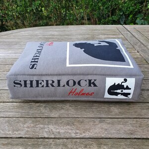 May include: A gray fabric book-shaped cushion with the text "SHERLOCK Holmes" printed on the front and side. The cushion features a silhouette of Sherlock Holmes in a white frame.