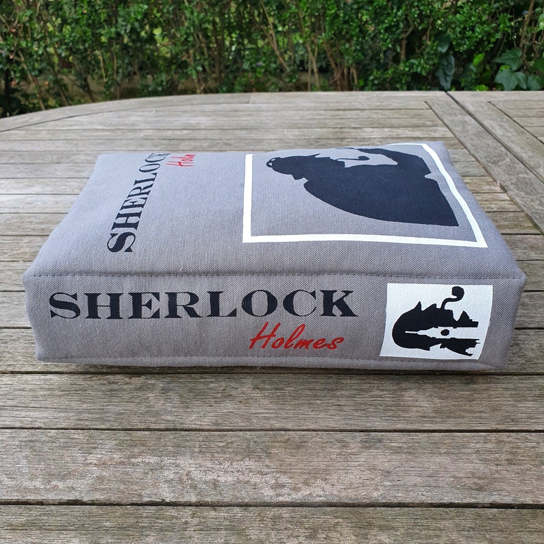 May include: A gray fabric book-shaped cushion with the text "SHERLOCK Holmes" printed on the front and side. The cushion features a silhouette of Sherlock Holmes in a white frame.