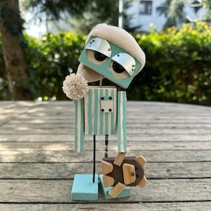 May include: A wooden robot with a blue and white striped body, a white hat, and large blue eyes. The robot is holding a small wooden bear in its hand.