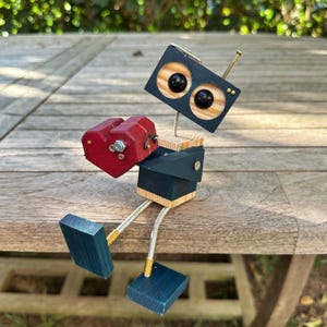 Może przedstawiać: Drewniany robot trzymający czerwone serce. Robot ma niebieskie i drewniane ciało z czarnymi oczami. Robot siedzi na drewnianej powierzchni.