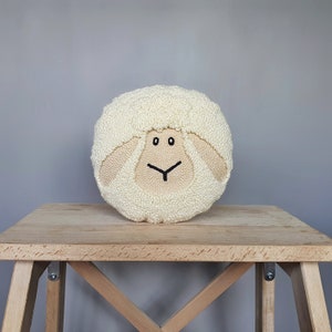 May include: A white crocheted sheep pillow with black embroidered eyes and a smiling mouth. The pillow is sitting on a wooden table.