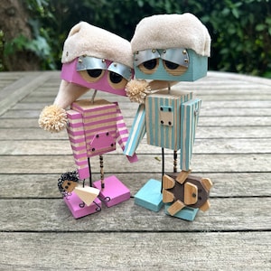May include: Two wooden robot figurines wearing white sleep caps. The robot on the left is pink with white stripes and holds a small pink figurine. The robot on the right is blue with white stripes and holds a brown wooden animal figurine.