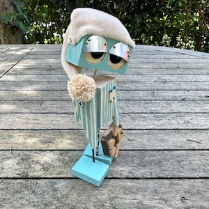 May include: A blue and white striped wooden robot wearing a white sleep cap and holding a wooden object. The robot is standing on a wooden surface.