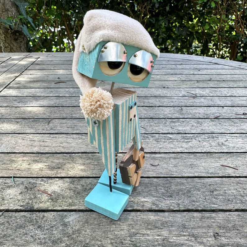 May include: A blue and white striped wooden robot wearing a white sleep cap and holding a wooden object. The robot is standing on a wooden surface.