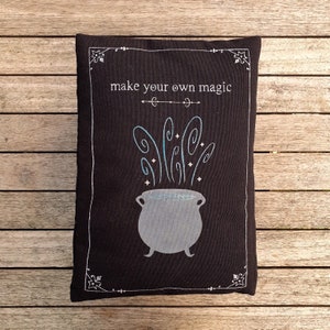 May include: Black pillow with white embroidery. The embroidery depicts a cauldron with steam rising from it and the text "make your own magic".