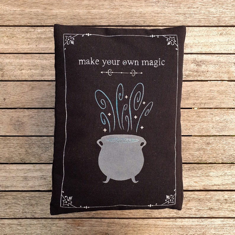 May include: Black pillow with white embroidery. The embroidery depicts a cauldron with steam rising from it and the text "make your own magic".