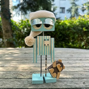 May include: A blue and white striped wooden robot wearing a white knit hat with a pom-pom. The robot has large, round, black eyes and is holding a small, brown wooden animal.